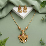 Gold-toned traditional jewelry set featuring a necklace with an ornate pendant and matching earrings, styled on a sage green draped fabric background with a white display block and small green leaves.
