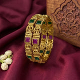 A gold-plated triple bangle bracelet with green and purple square stone accents, displayed on a draped dark red fabric next to white jasmine flowers and a carved wooden box.