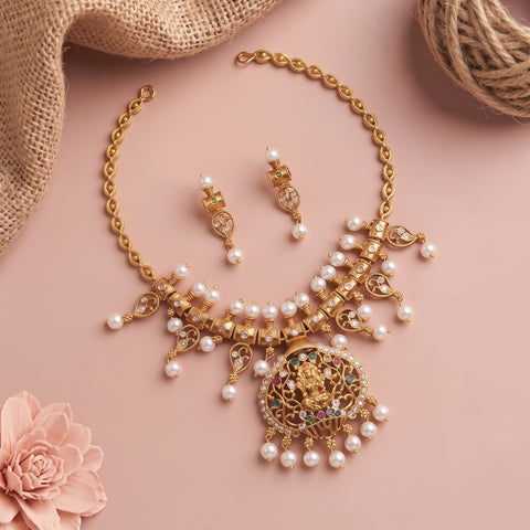 Gold-toned traditional necklace and matching earrings with white faux pearls and a central deity pendant, styled on a soft pink background with burlap and a pink flower prop.