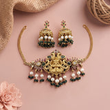 Gold-toned traditional jewelry set featuring a necklace with a central deity motif, dangling green stones and white pearl beads, matching drop earrings, styled on a soft pink background with burlap and a flower prop.