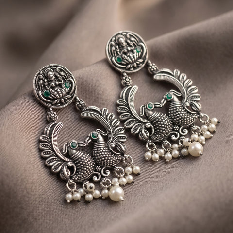 A pair of ornate silver-toned Indian-style dangle earrings with detailed filigree, small green stone accents, and clusters of white faux pearls, displayed on a draped brown fabric background.