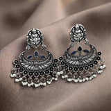 A pair of ornate silver-toned Indian jhumka earrings with black oxidized detailing, small blue stone accents, and dangling bead chains, displayed on a draped brown fabric background.