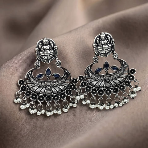 A pair of ornate silver-toned Indian jhumka earrings with black oxidized detailing, small blue stone accents, and dangling bead chains, displayed on a draped brown fabric background.