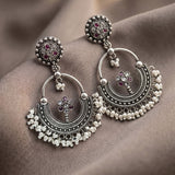 A pair of ornate silver-toned huggie earrings with floral stud tops, crescent-shaped dangling elements, and rows of small white pearl-like beads, displayed on draped taupe fabric.
