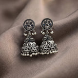 A pair of ornate silver-toned Indian jhumka earrings with detailed filigree, small dark blue stones, and dangling white pearl beads, displayed on draped taupe fabric.