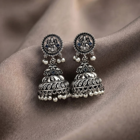 A pair of ornate silver-toned Indian jhumka earrings with detailed filigree, small dark blue stones, and dangling white pearl beads, displayed on draped taupe fabric.