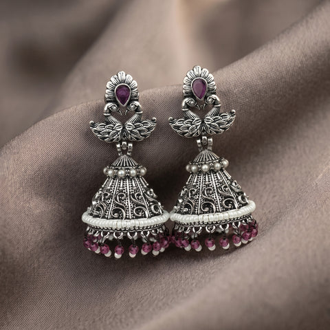 A pair of ornate silver-toned jhumka earrings with purple gemstone accents, displayed on draped brown fabric.