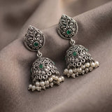 A pair of ornate silver-toned Indian jhumka earrings with green stone accents and dangling pearl-like beads, displayed on draped taupe fabric.