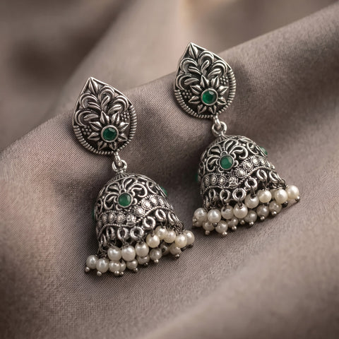 A pair of ornate silver-toned Indian jhumka earrings with green stone accents and dangling pearl-like beads, displayed on draped taupe fabric.