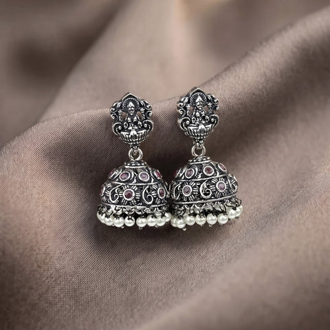 A pair of traditional Indian silver-toned jhumka earrings with intricate filigree work, small pink stone accents, and rows of white pearl beads at the bottom, displayed on a draped taupe fabric background.
