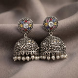 A pair of ornate silver-toned Indian jhumka earrings with multicolored stone accents and dangling pearl-like bells, displayed on draped taupe fabric.
