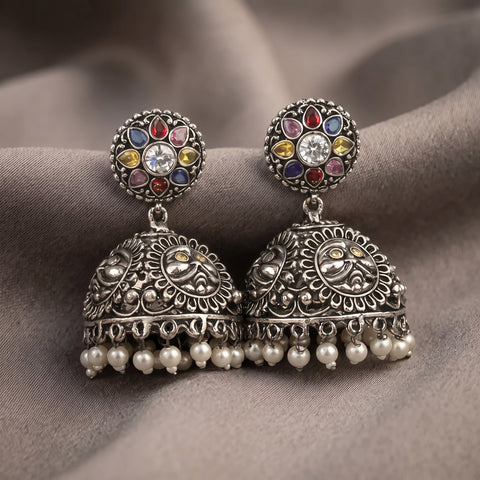 A pair of ornate silver-toned Indian jhumka earrings with multicolored stone accents and dangling pearl-like bells, displayed on draped taupe fabric.