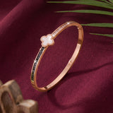 A rose gold bangle bracelet with a white mother-of-pearl four-leaf clover charm, set against a dark burgundy fabric background with decorative green leaves.