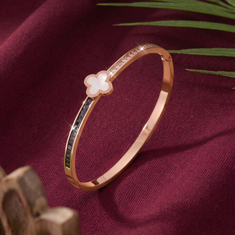 A rose gold bangle bracelet with a white mother-of-pearl four-leaf clover charm, set against a dark burgundy fabric background with decorative green leaves.
