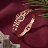 Rose gold bangle bracelet with a circular interlocking design and a bezel-set crystal accent, displayed on a deep burgundy fabric background with decorative leaves and wood.