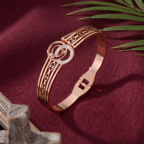 Rose gold bangle bracelet with a circular interlocking design and a bezel-set crystal accent, displayed on a deep burgundy fabric background with decorative leaves and wood.