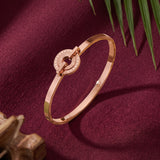 A rose gold bangle bracelet with a circular open-center clasp, styled on a textured dark burgundy fabric background with green leaves and a wooden prop.