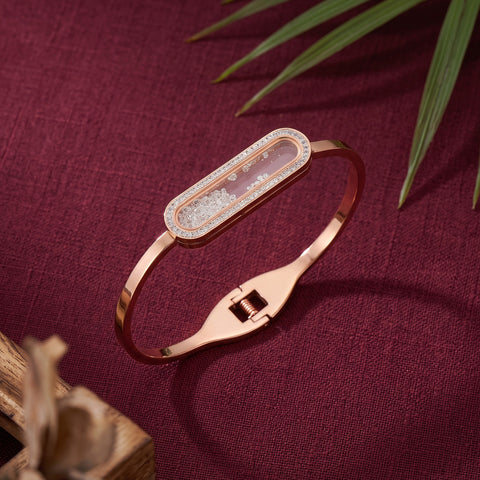 A rose gold bangle bracelet with a rectangular clear crystal-encrusted bezel and a central white textured inlay, displayed on a textured burgundy fabric background with decorative green leaves.