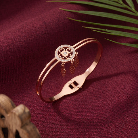 A rose gold bangle bracelet with a circular floral motif and hanging leaf charms, displayed on a textured burgundy fabric background with decorative green leaves and a wooden object.