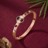 A rose gold bangle bracelet with a central four-petal motif in black and white enamel, accented with small black and white teardrop inlays, displayed on a textured burgundy fabric background with green leaves.