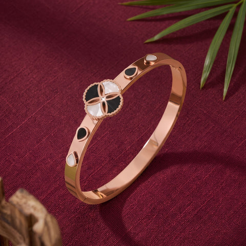 A rose gold bangle bracelet with a central four-petal motif in black and white enamel, accented with small black and white teardrop inlays, displayed on a textured burgundy fabric background with green leaves.