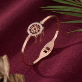A rose gold bangle bracelet with a circular filigree flower charm and small dangling charms, displayed on a textured burgundy fabric background with decorative palm leaves.