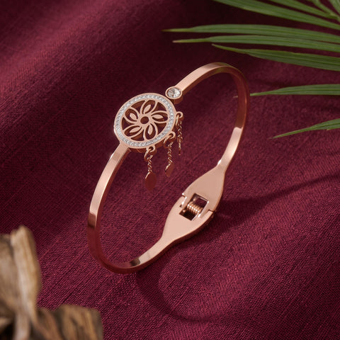 A rose gold bangle bracelet with a circular filigree flower charm and small dangling charms, displayed on a textured burgundy fabric background with decorative palm leaves.