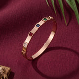 A rose gold bangle bracelet with four square bezel-set gemstone accents in red, green, blue, and clear, displayed on a textured burgundy fabric background with green leaves.