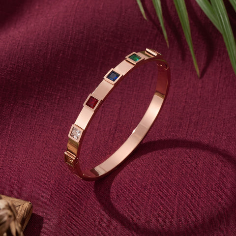 A rose gold bangle bracelet with four square bezel-set gemstone accents in red, green, blue, and clear, displayed on a textured burgundy fabric background with green leaves.