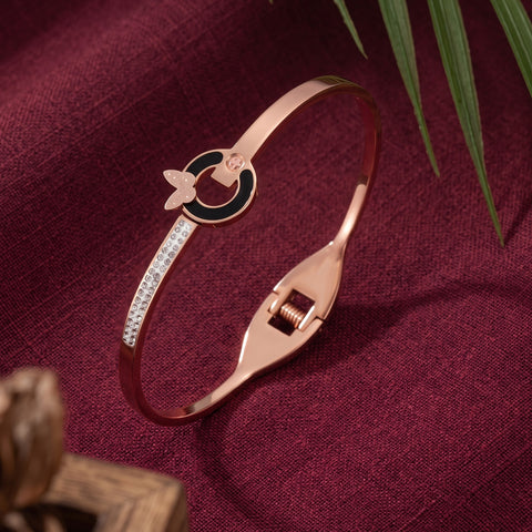 A rose gold bangle bracelet with a black enamel circle, a butterfly motif, and a row of clear crystals, displayed on a textured burgundy fabric background with green leaves.