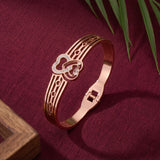 A rose gold bangle bracelet with a heart-shaped design and small clear rhinestones, displayed on a textured dark red fabric background with decorative green leaves and a wooden corner.