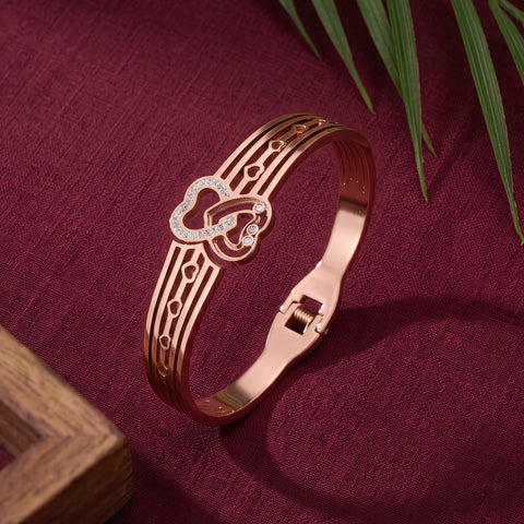 A rose gold bangle bracelet with a heart-shaped design and small clear rhinestones, displayed on a textured dark red fabric background with decorative green leaves and a wooden corner.