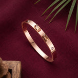 A rose gold bangle bracelet with rectangular baguette-cut clear crystals and small round clear stones, displayed on a textured burgundy fabric background with decorative green leaves and a wooden corner.