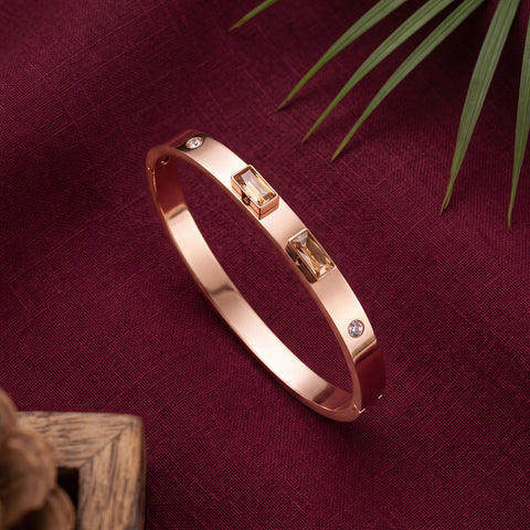 A rose gold bangle bracelet with rectangular baguette-cut clear crystals and small round clear stones, displayed on a textured burgundy fabric background with decorative green leaves and a wooden corner.