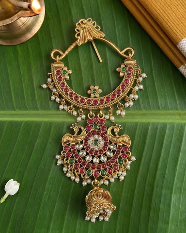 An ornate gold-toned traditional Indian jewelry piece, likely a necklace or pendant, decorated with red and green stones and white pearl beads, displayed on a green banana leaf.