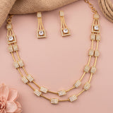 Gold-plated double-strand necklace and matching drop earrings with square crystal-studded accents, arranged on a soft pink background with burlap and a pink flower prop.