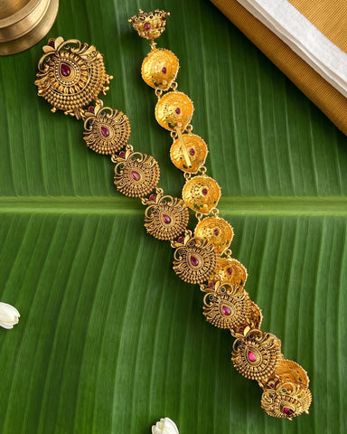 An ornate gold-toned traditional jewelry set with matching earrings and a necklace, featuring circular motifs and small red gemstone accents, displayed on a large green banana leaf.