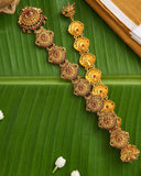 A gold-toned traditional Indian jewelry set, including a necklace and matching earrings, decorated with red and green stones, displayed on a green banana leaf.