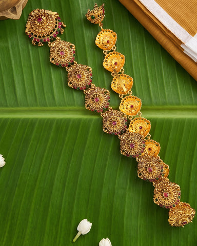 A gold-toned traditional Indian jewelry set, including a necklace and matching earrings, decorated with red and green stones, displayed on a green banana leaf.