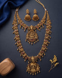 An ornate gold-toned traditional jewelry set featuring a necklace, matching earrings, and a central pendant with detailed filigree, red stone accents, and dangling beads, arranged on a dark navy background with decorative fabric and small dried flowers.