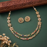 Gold-toned floral jewelry set with a necklace and matching earrings, decorated with multicolored stones, displayed on a dark green background with a carved wooden prop and small dried flowers.