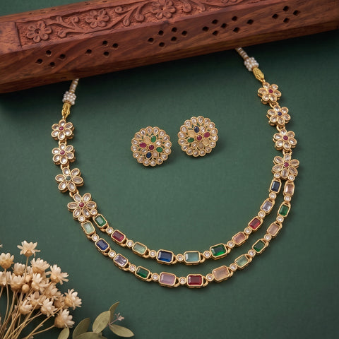 Gold-toned floral jewelry set with a necklace and matching earrings, decorated with multicolored stones, displayed on a dark green background with a carved wooden prop and small dried flowers.