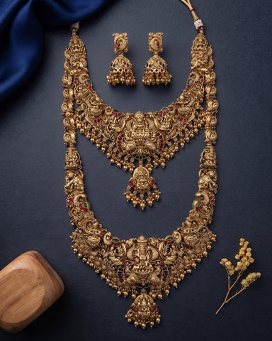 An ornate gold-toned traditional necklace set with matching earrings, arranged on a dark navy background with decorative blue fabric, a wooden block, and small dried flowers.