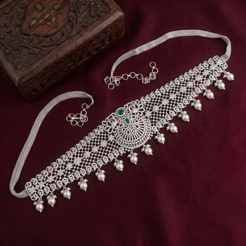 A silver-toned traditional Indian choker necklace with intricate filigree work, hanging pearl-like beads, and a central green stone, displayed on a deep burgundy fabric next to a carved wooden box.