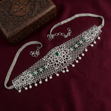 A silver-toned Indian choker necklace with intricate filigree cutouts, hanging silver beads, and green stone accents, displayed on a deep maroon fabric background next to a carved wooden box.