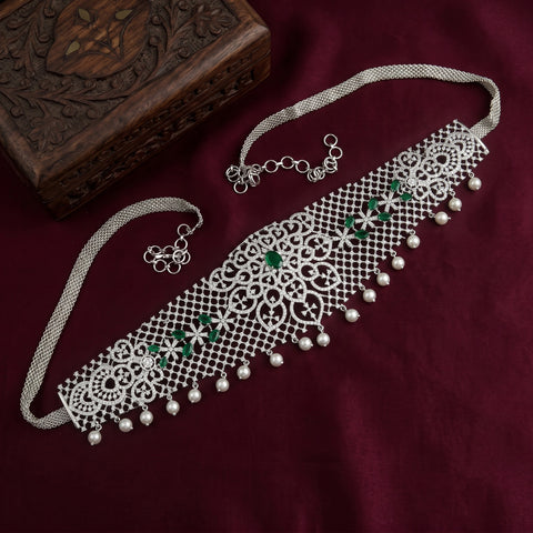 A silver-toned Indian choker necklace with intricate filigree cutouts, hanging silver beads, and green stone accents, displayed on a deep maroon fabric background next to a carved wooden box.