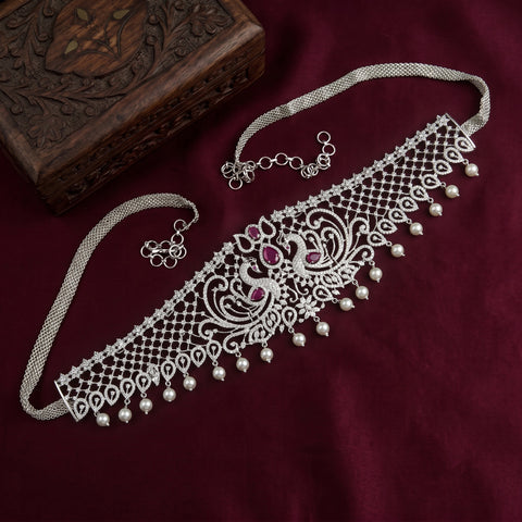A silver-toned traditional Indian choker necklace with intricate filigree work, small pearl-like beads, and two pink gemstone accents, displayed on a dark burgundy fabric next to a carved wooden box.