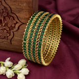 A set of five gold-plated bangles decorated with green rectangular stones and gold chain links, displayed on a deep burgundy fabric next to a carved wooden box and white jasmine flowers.
