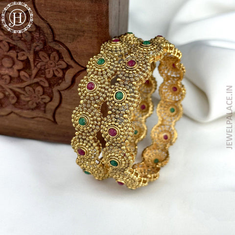 Two gold-plated bangle bracelets with vibrant Kemp stones, displayed against a wooden backdrop with intricate gold patterns.