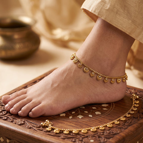 A close-up of a person's bare foot wearing a delicate gold anklet with crescent moon and floral motifs, standing on a carved wooden tray with a matching gold anklet laid out nearby.